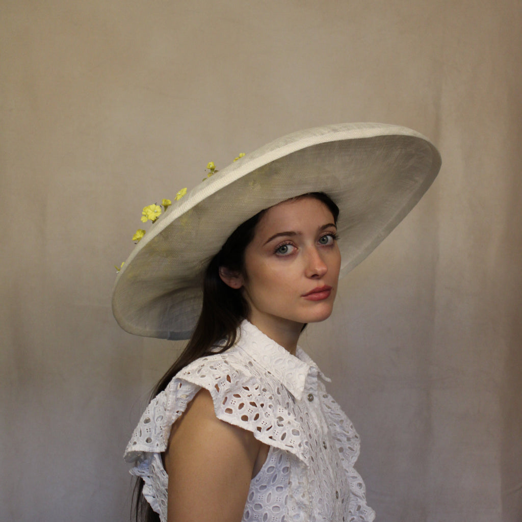 A woman wearing a cream and yellow wide-brimmed hat with vintage flowers, paired with a white lace top. Cream and yellow hat with vintage flowers and silk abaca