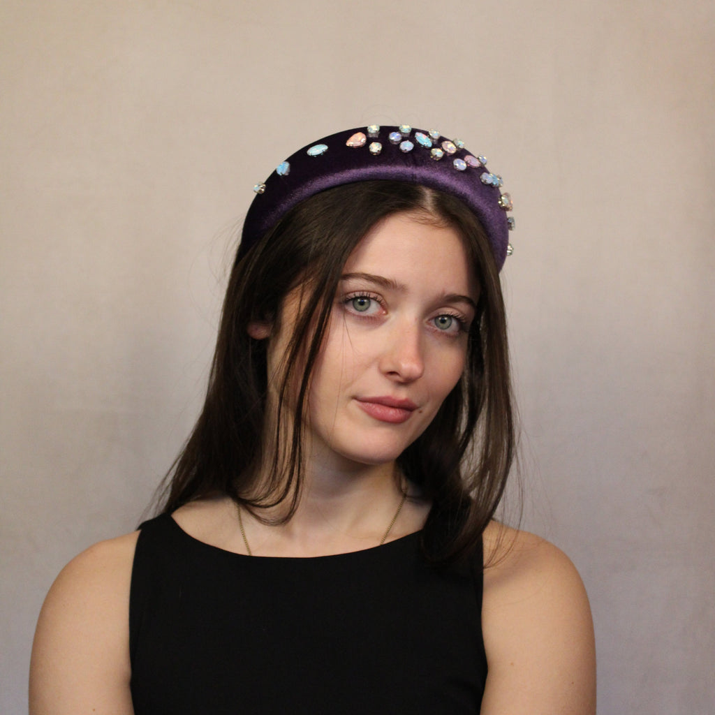 A person wearing a purple velour headband with pastel colored gemstones on it. Purple velour headband set with pastel gemstones