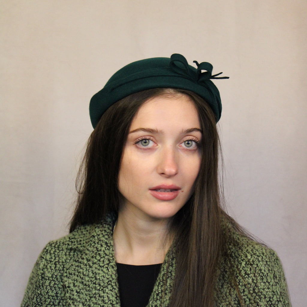 A woman wearing a bottle green wool hat with detailed trim. Bottle green wool hat with detailed trim.